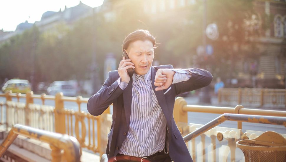 photo of man in blue blazer and striped shirt standing by stairs while on the phone and looking at his watch