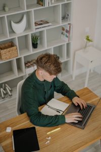 high angle shot of a male student in green long sleeves using a laptop on a wooden desk