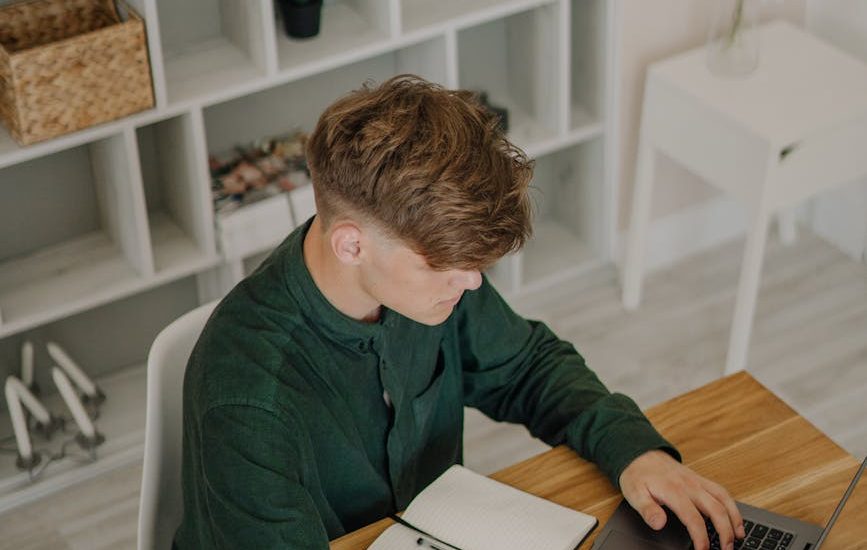 high angle shot of a male student in green long sleeves using a laptop on a wooden desk