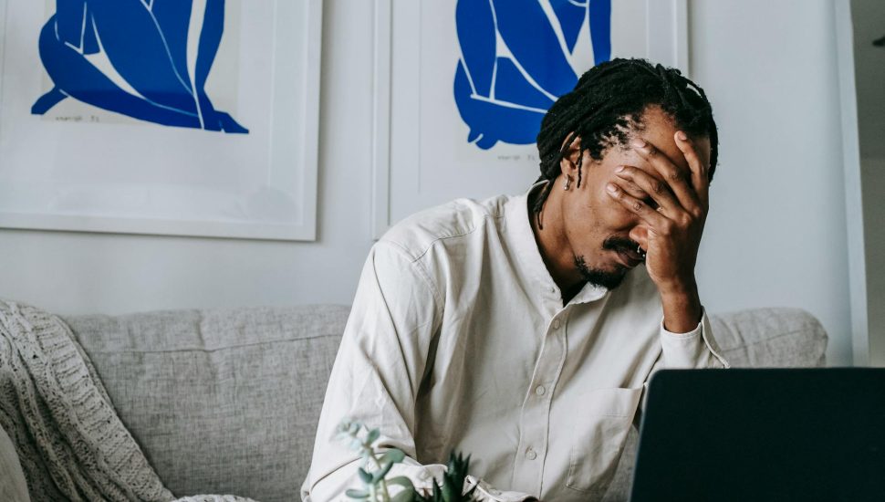 upset young black guy covering face with hand while working remotely on netbook
