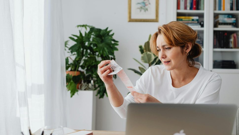 woman sitting behind a desk using laptop and looking at items on a receipt